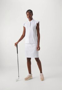 Egy nő áll egy golfütővel, fehér ujjatlan pólót, fehér szoknyát és világos rózsaszín sportcipőt visel, egy sima háttér előtt.