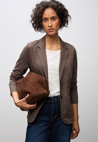 Blazer marrone con una texture morbida, abbinato a una t-shirt bianca e jeans scuri. Tiene in mano una borsa in suede marrone con una forma strutturata.