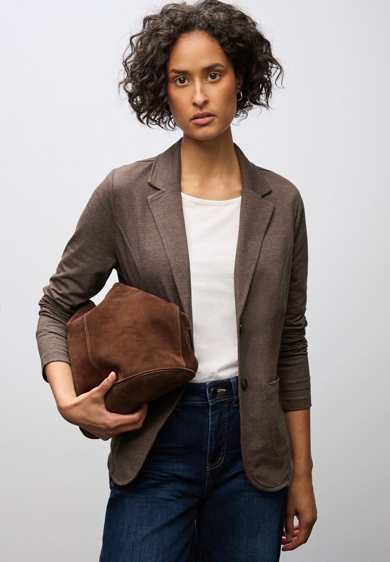 Blazer marrone con una texture morbida, abbinato a una t-shirt bianca e jeans scuri. Tiene in mano una borsa in suede marrone con una forma strutturata.