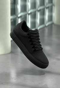 Μαύρο παπούτσι σπορ χαμηλού προφίλ με παχιά σόλα και μαύρα κορδόνια, αιωρούμενο πάνω από ένα λείο γκρι πάτωμα με φόντο έναν τοίχο από γυάλινα μπλοκ.