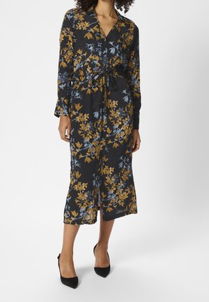 Femme portant une robe midi noire avec imprimé floral jaune et bleu, manches longues, boutonnée sur le devant, ceinture à la taille, et talons pointus noirs.