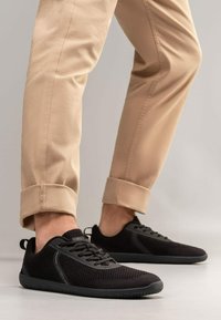 Black mesh sneakers with textured fabric, lace-up design, and rubber soles. Worn with rolled beige chinos, showcasing a sleek silhouette.