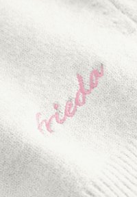 Witte stof met een zachte textuur heeft roze geborduurde tekst die "frieda" leest. Het ontwerp is eenvoudig en strak.