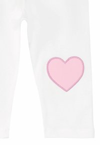 Valkoiset legginsit, joissa on suuri vaaleanpunainen brodeerattu sydänkuvio vasemmalla puolella. Pehmeä kangas ja tiukka muoto. Samettimainen pinta sydämessä.