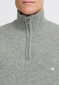 Maglione grigio a coste con colletto alto e chiusura con zip. Presenta un piccolo logo bianco sul petto, realizzato in materiale lavorato a maglia.
