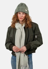Groene gebreide beanie, olijfgroen bomberjack, lichtgrijze gestreepte sjaal en blauwe spijkerbroek. Gelaagd breipatroon en zichtbare hardwaredetails.