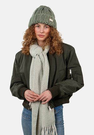 Bonnet tricoté vert, bomber olive, écharpe rayée gris clair et jean bleu. Motif de tricot texturé et détails matériels visibles.
