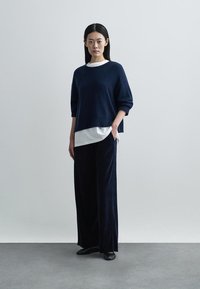 Tumši zils adīts džemperis ar vaļīgu piegriezumu, kopā valkāts ar baltu kārtainu topu un plašām melnām biksēm no mīksta, teksturēta auduma. Melni apavi.