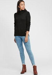 Schwarzer Strickpullover mit hohem Kragen, gerippten Bündchen und Saum. Kombiniert mit hellblauen Jeans und braunen Stiefeletten mit fransigen Kanten.