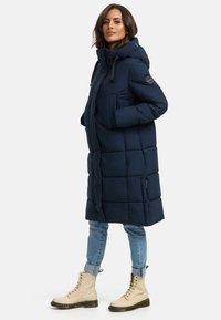 Navy blue padded coat with a hood, featuring a quilted design, front pockets, and a zipper. Paired with light jeans and beige lace-up boots.