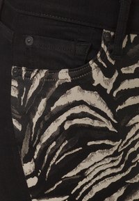 Jeans negros con un patrón de rayas de cebra en beige claro en el bolsillo delantero y el muslo superior, con botones metálicos estándar y una tela resistente.