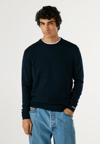 Sudadera de cuello redondo en azul marino de tela suave, con puños y dobladillo acanalados. Combinada con jeans ajustados de color azul claro.