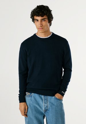 Navyblaues Rundhals-Sweatshirt aus weichem Material mit gerippten Bündchen und Saum. Getragen mit hellblauen taillierten Jeans.
