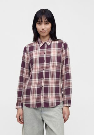 Femme portant une chemise à manches longues à carreaux violets et roses boutonnée, avec un pantalon gris clair, sur un fond blanc uni.