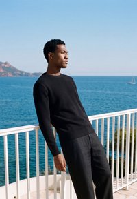 Jeune homme en pull noir appuyé sur une rambarde blanche avec une mer calme et un voilier lointain en arrière-plan sous un ciel bleu clair.
