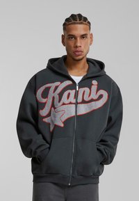 Mørkegrå zip-up hoodie med et fremtrædende rødt og sølvbroderet "Kani"-logo og stjernedesign. Har snøre i hætten og lommer foran.