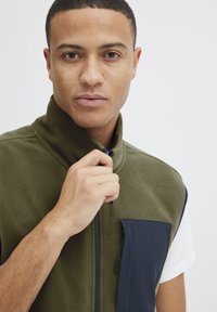 Grønn fleece vest med høy krage, marineblå lomme og kontrastfarge i marineblått. Glatt tekstur og glidelås foran.