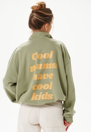 Jeune femme aux cheveux relevés en chignon portant un sweat-shirt vert olive avec l'inscription « Cool mums have cool kids » imprimée dans le dos en gros caractères jaunes.