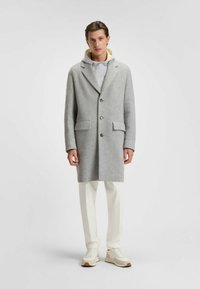 Cappotto grigio in lana con colletto a reversing, chiusura a un petto, due tasche frontali, indossato sopra un hoodie grigio chiaro e pantaloni bianchi.