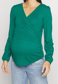 Donna incinta che indossa un top avvolto a maniche lunghe color turchese e jeans azzurri chiari, in piedi contro uno sfondo chiaro e uniforme.