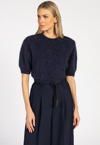 Vrouw met blond haar, gekleed in een navy textuur short-sleeve sweater en hooggetailleerde navy broek met een gevlochten riem, die iets naar links kijkt.