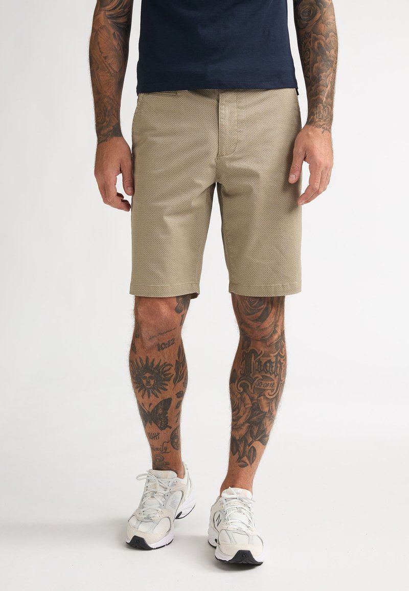 Homme portant un short texturé beige, des baskets blanches, une chemise bleu marine, avec des bras et des jambes fortement tatoués, debout sur un fond blanc.