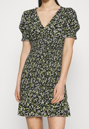 Kvinde iført sort blomsterprint kortærmet kjole med V-hals, smocket talje og flæsekant, der står mod en hvid baggrund.