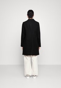 Cappotto di lana nero con maniche lunghe e colletto, vestibilità aderente, che arriva a metà coscia. Abbinato a pantaloni larghi crema e sneakers.