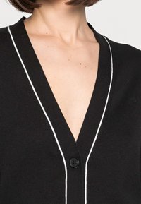 Svart cardigan med vit kant, V-ringning och enstaka knappstängning. Mjuk textur och figurnära design som framhäver kragen.