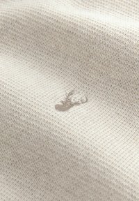 Tejido de punto beige con un pequeño logotipo de cabeza de ciervo bordado en hilo marrón claro cerca del centro.
