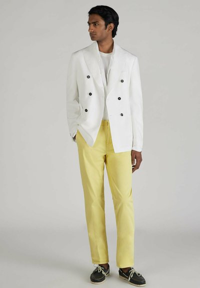 Blazer bianco doppiopetto con bottoni neri, abbinato a pantaloni giallo chiaro e scarpe scure con lacci. Sotto una semplice camicia bianca a girocollo.