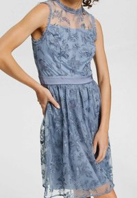 Vestido azul bordado con escote transparente, patrones florales y una cinta en la cintura. La tela es suave y tiene un diseño fluido.