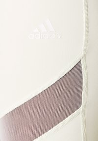Bílé sportovní legíny s nenápadným logem Adidas, které mají texturovanou hnědou síťovinovou část a jemné švy.