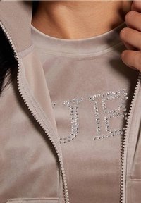 Beige velours zip-up jas met een zachte textuur, gelaagd over een bijpassend top met strass letterdetails.