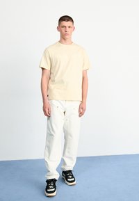Beige bomulds t-shirt med "Timberland" logo, parret med hvide baggy bukser med guld accenter, og sorte sneakers med hvide striber.