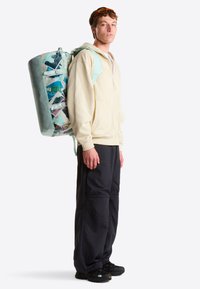 Mochila grande y colorida con un diseño gráfico, correas acolchadas y un acento turquesa. Se lleva con una sudadera con capucha crema con cremallera y pantalones negros.