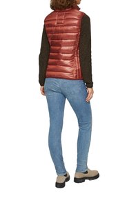 Rote gepolsterte Weste mit hohem Kragen und Nähten; kombiniert mit einem dunklen Pullover und blauen Jeans. Die Schuhe sind hellbraun mit einer dicken Sohle.