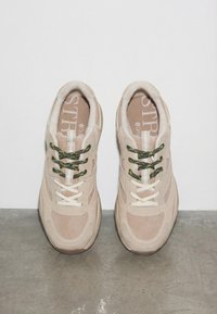 Baskets en daim beige avec semelles blanches et lacets dépareillés, une paire noire et verte, l'autre blanche, posées sur un sol en béton.