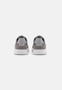 Šedé a námořnické tenisky s hladkým semišovým povrchem, polstrovanou podporou kolem kotníku a bílými gumovými podrážkami. Minimalistický design, pohled zpoza.