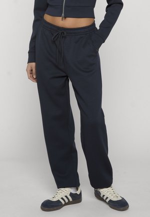Personne portant un pantalon de jogging bleu marine à cordon, une veste zippée courte bleu marine et des baskets bleu marine avec des rayures blanches et des lacets, debout avec une main dans la poche.