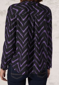 Blouse noire avec un motif en zigzag violet. Manches longues, fermeture à boutons dans le dos, coupe droite et texture de tissu douce et lisse.