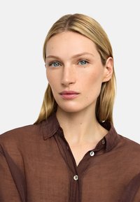 Brązowa, przezroczysta koszula na guziki z kołnierzykiem, o subtelnej teksturze i widocznym przeszyciu. Model ma blond włosy i niebieskie oczy.