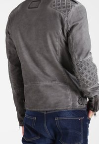 Giacca da motociclista in pelle grigia con pannelli imbottiti sulle spalle, una finitura testurizzata e cerniere laterali. Cuciture visibili e design aderente.