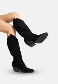 Schwarze kniehohe Stiefel aus Wildleder mit bestickten Details, spitz zulaufender Zehenpartie und Blockabsatz. Reißverschluss an der Seite.