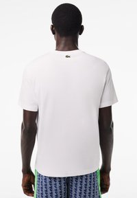 Camiseta de algodón blanca de mangas cortas, con un pequeño logo verde en la nuca. Textura suave, diseño clásico de cuello redondo.