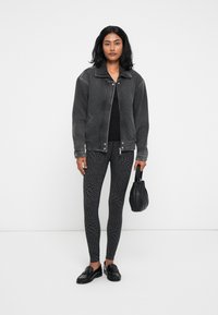 Svart denimjakke, svart tettsittende topp, mønstrede svarte leggings og svarte loafers. Modell som holder en liten svart veske, står mot en nøytral bakgrunn.