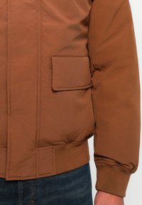 Brązowa kurtka puchowa z ribbowanym dołem i mankietami, boczną kieszenią oraz gładkim, teksturowanym wykończeniem. Luźny, casualowy fason z lekko oversize'owym krojem.