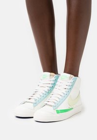 Höga sneakers i vitt läder med ljusblå akcentpaneler, grön swoosh och texturerade detaljer. Snören i vitt.