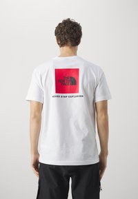 Hvit bomulls T-skjorte med korte ermer. Har et rødt kvadratisk grafikk på baksiden med svart tekst "THE NORTH FACE" og "NEVER STOP EXPLORING."