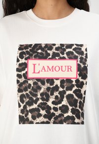 Biele tričko s dizajnom štvorca v leopardom vzore a ružovou obdĺžnikovou etiketou s nápisom "L'amour" v tučných písmenách.